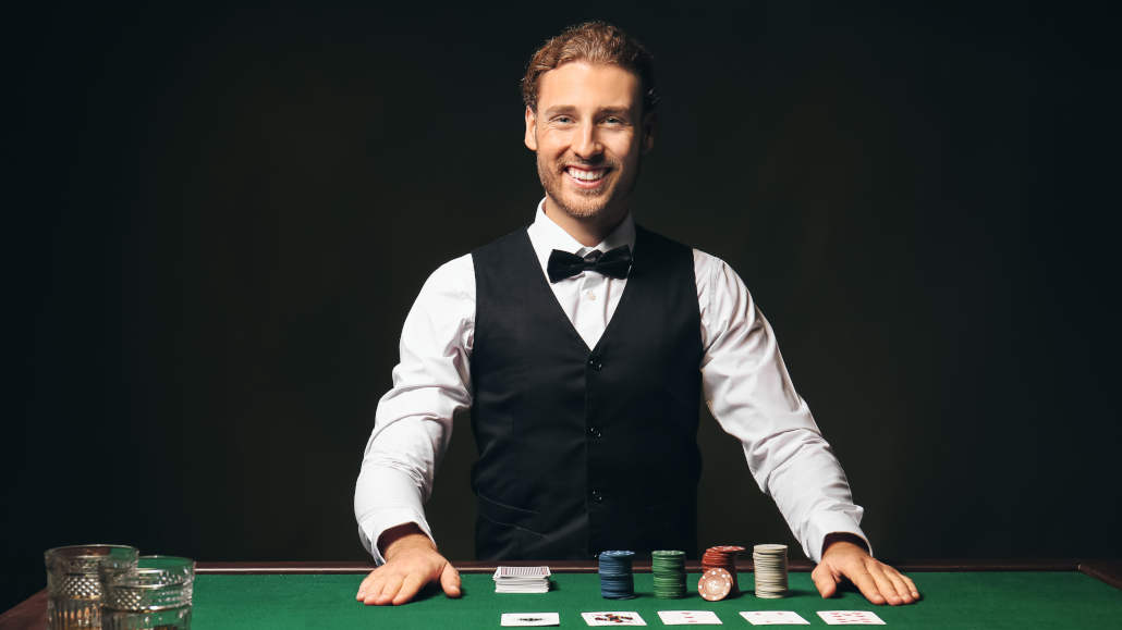 A smiling professional dealer standing behind a green felt poker table with cards and chips